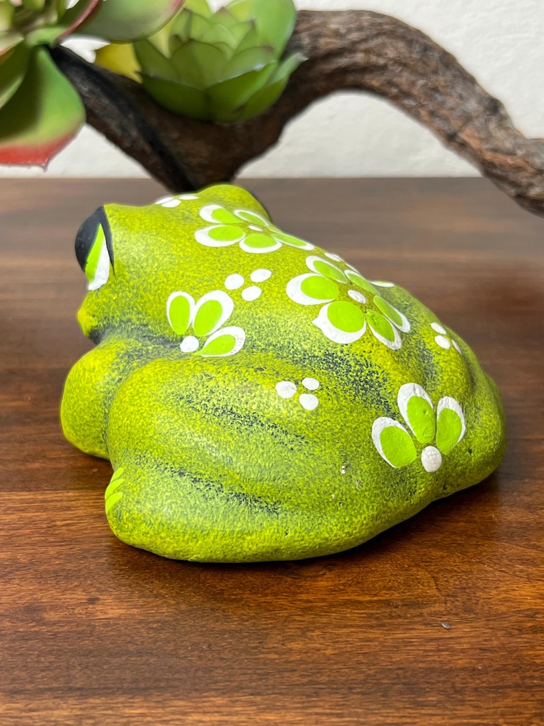May include: A ceramic frog figurine in a vibrant green hue, adorned with white and lime green floral patterns. The frog has black eyes and is positioned on a wooden surface. A decorative item for home decor.