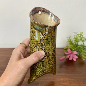 May include: A wall-mounted ceramic vase featuring a textured olive green glaze with a dark crackle pattern and intricate lace-like impressions. The vessel has a wide, slanted opening with a small circular hole at the top for hanging purposes.
