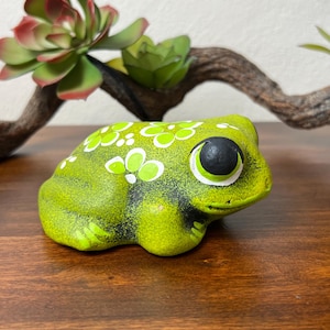 May include: A ceramic frog figurine in shades of green with white floral accents. The frog has large, black eyes and is positioned on a wooden surface. Decorative succulent plants and a branch are in the background.