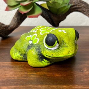 May include: A ceramic frog figurine in shades of green and black, with white floral accents. The frog has large, expressive eyes and is positioned on a wooden surface. Decorative succulents and a branch are in the background.