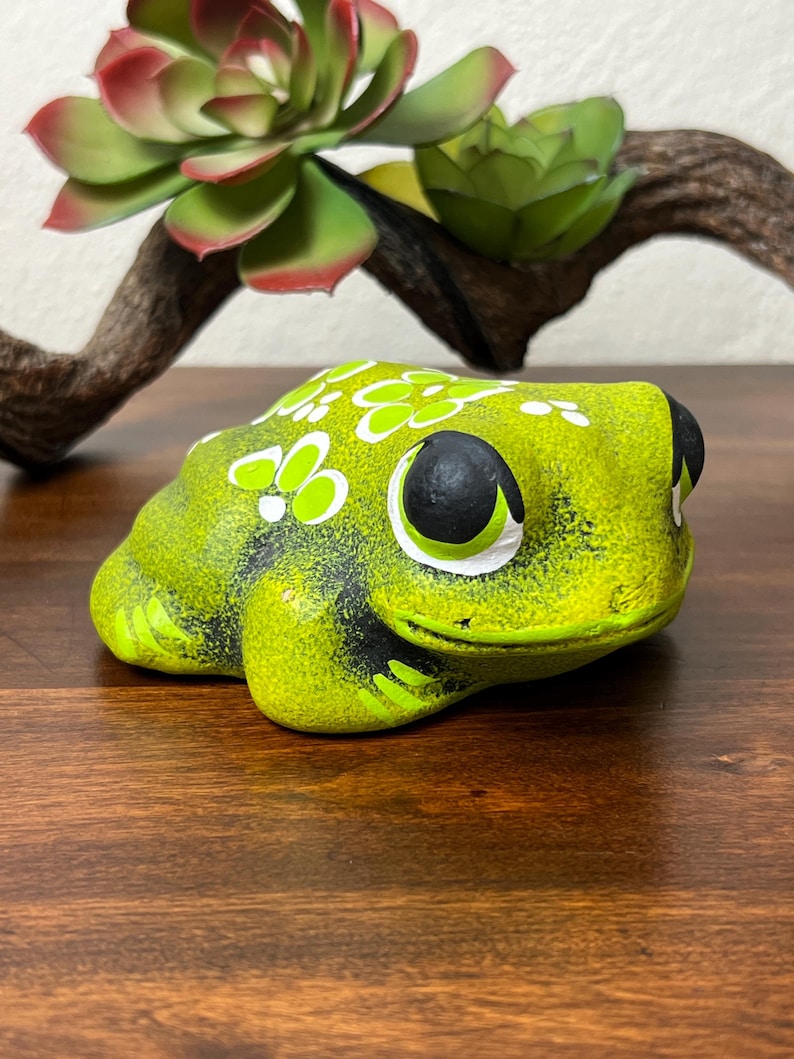 May include: A ceramic frog figurine in shades of green and black, with white floral accents. The frog has large, expressive eyes and is positioned on a wooden surface. Decorative succulents and a branch are in the background.