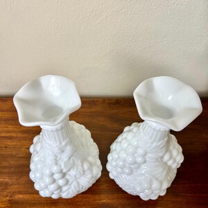 Puede incluir: Dos jarrones de cristal blancos de estilo vintage. Cada jarr&oacute;n presenta una base bulbosa decorada con un dise&ntilde;o de racimo de uvas y una parte superior acampanada y con volantes. Los jarrones se exhiben sobre una superficie de madera.