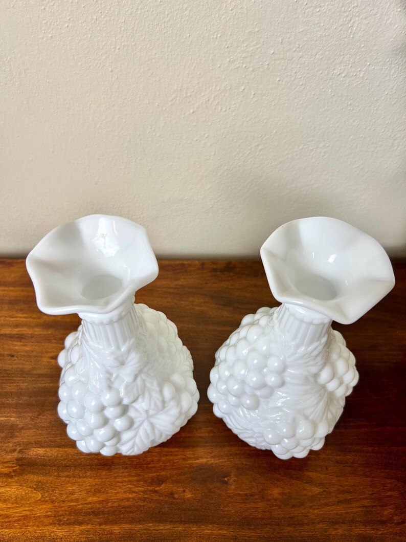 Puede incluir: Dos jarrones de cristal blancos de estilo vintage. Cada jarr&oacute;n presenta una base bulbosa decorada con un dise&ntilde;o de racimo de uvas y una parte superior acampanada y con volantes. Los jarrones se exhiben sobre una superficie de madera.