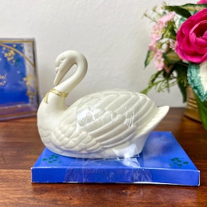 Może przedstawiać: Biała ceramiczna figurka łabędzia ze złotą opaską na szyi, spoczywająca na niebieskim pudełku. Łabędź ma szczegółowe tekstury piór. W tle bukiet różowych kwiatów i oprawiony obraz.