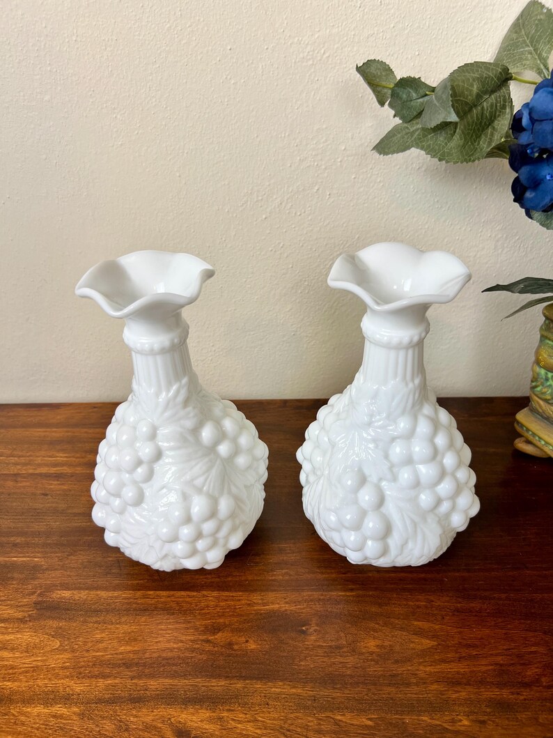 Puede incluir: Dos jarrones blancos de estilo vintage. Cada jarr&oacute;n presenta una parte superior con volantes, un cuello acanalado y un cuerpo con forma de racimo de uvas. Los jarrones son de vidrio blanco opaco y est&aacute;n sobre una superficie de madera oscura.