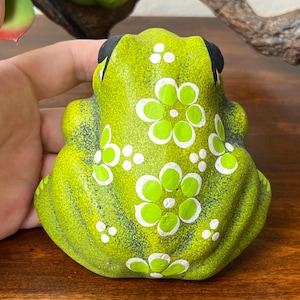 May include: A lime green ceramic frog figurine with white floral accents and black eyes. The frog is decorated with white dots and white flower designs. The figurine is sitting on a wooden surface.