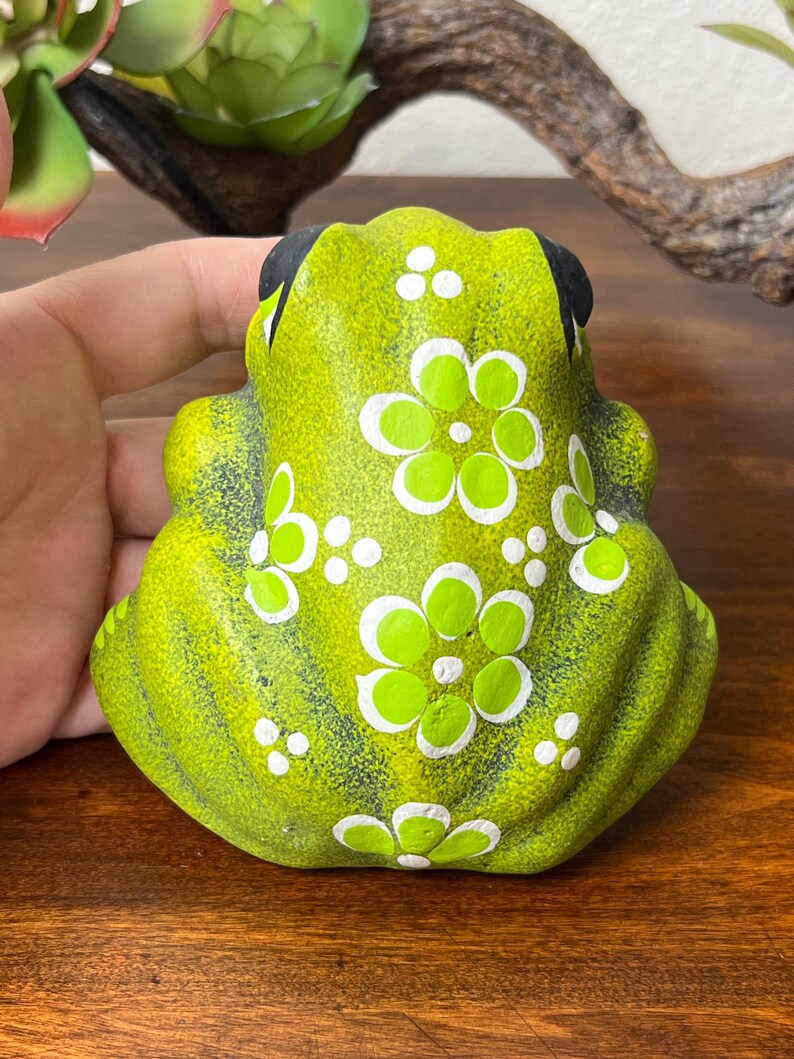 May include: A lime green ceramic frog figurine with white floral accents and black eyes. The frog is decorated with white dots and white flower designs. The figurine is sitting on a wooden surface.