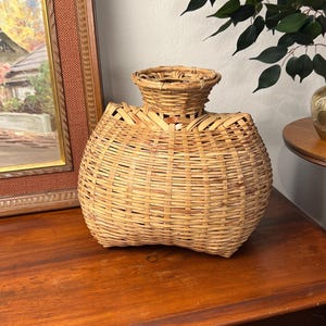 May include: A woven basket with a unique shape, featuring a wide base and a narrower neck. The basket is made of light brown, natural-colored reeds, and is displayed on a wooden surface. The basket's design suggests it could be used for decorative purposes or storage.