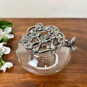 May include: A clear glass bowl with a decorative silver-coloured lid featuring a hummingbird and floral design. The bowl sits on a dark wooden surface, with white flowers and green leaves in the background. The lid is intricately detailed.