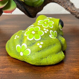 May include: A lime green ceramic frog figurine with white and light green floral designs. The frog has black eyes and is sitting on a wooden surface. Decorative item for home decor.