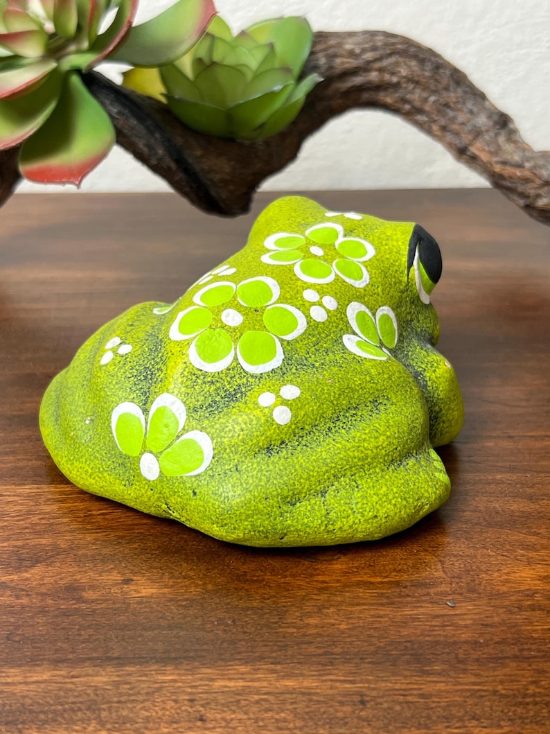 May include: A lime green ceramic frog figurine with white and light green floral designs. The frog has black eyes and is sitting on a wooden surface. Decorative item for home decor.