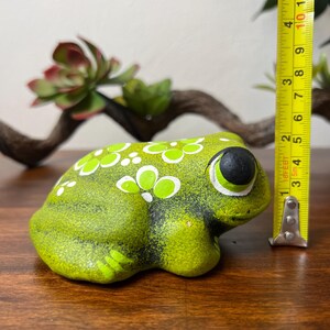 May include: A green ceramic frog figurine with white and light green floral accents. The frog has large black eyes with white outlines. The figurine is approximately 3 inches long.