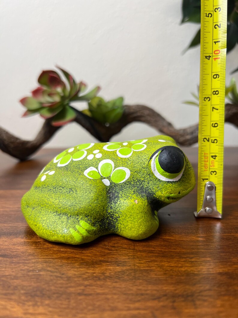 May include: A green ceramic frog figurine with white and light green floral accents. The frog has large black eyes with white outlines. The figurine is approximately 3 inches long.