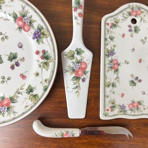 May include: A white ceramic plate, cake server, cheese knife, and small cutting board with a fruit and floral design. The design includes peaches, grapes, and green leaves. The plate has a gold rim.