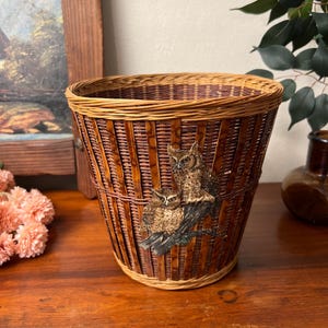 May include: A woven wicker wastebasket with a brown and tan color scheme. The wastebasket features a painted image of two owls perched on a branch. The basket has a flared shape and a woven rim.