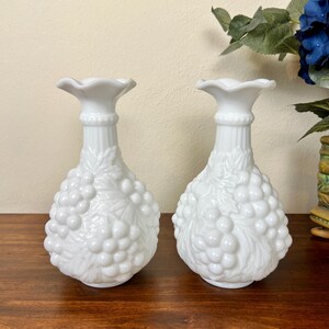 Puede incluir: Dos jarrones blancos de cristal de leche. Cada jarr&oacute;n tiene una parte superior con volantes, un cuello acanalado y un cuerpo con forma de racimo de uvas. Los jarrones est&aacute;n sobre una superficie de madera oscura.