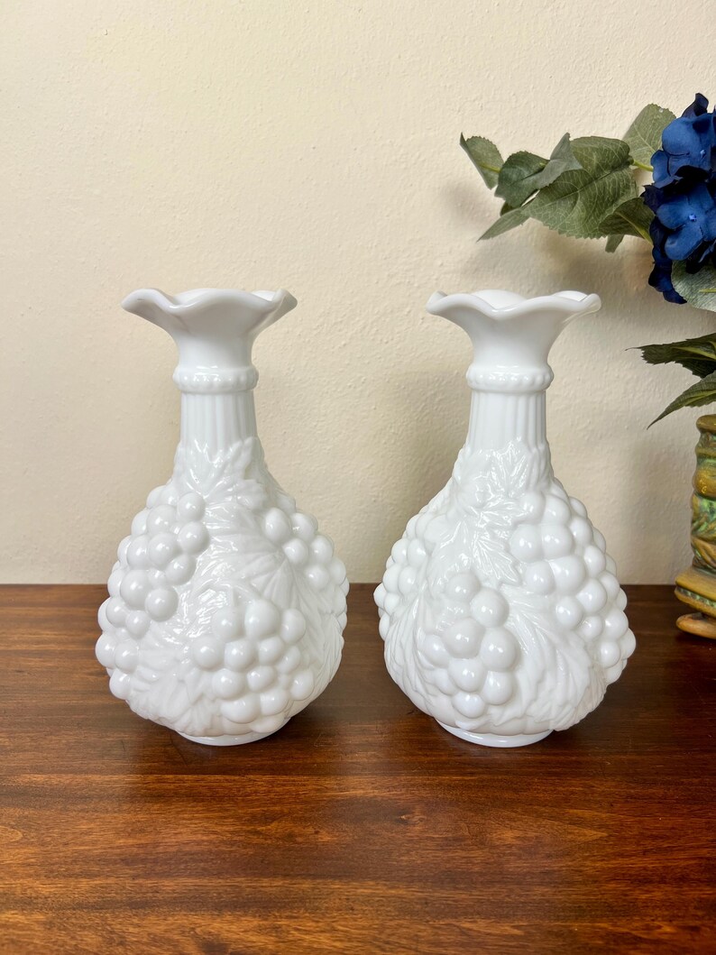 Puede incluir: Dos jarrones blancos de cristal de leche. Cada jarr&oacute;n tiene una parte superior con volantes, un cuello acanalado y un cuerpo con forma de racimo de uvas. Los jarrones est&aacute;n sobre una superficie de madera oscura.