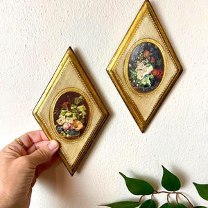 May include: Two diamond-shaped wall art pieces with gold frames and oval floral paintings. The frames have a textured, cream-coloured background. One is held by a hand. The paintings feature colourful flowers.