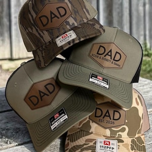 May include: A stack of Richardson 112PFP trucker hats with a brown leatherette patch that says "DAD". Some hats have camouflage patterns, and others are olive green. One hat says "BODIE & EVERETT & EMMA" and another says "EST. 2026".