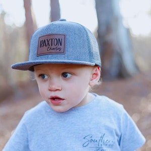 May include: A gray trucker hat with a brown leather patch that reads "Raxton Charles." The hat has a mesh back and a flat brim. The child is also wearing a gray t-shirt with the text "Southern" and "Est. 2014."