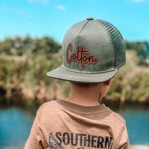 May include: A young person wearing a brown t-shirt with a duck hunting graphic and the text "Southern Refinery Duck Hunting". The person is also wearing a green mesh trucker hat with the word "Cotton" in brown letters.