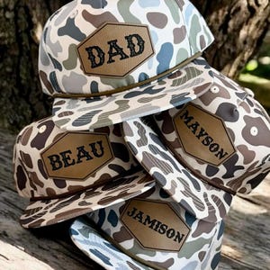 May include: A stack of camouflage baseball caps with brown leather patches. The patches display the names "DAD", "BEAU", "MAYSON", and "JAMISON" in black lettering. The caps feature a mix of white, blue, brown, and green patterns.