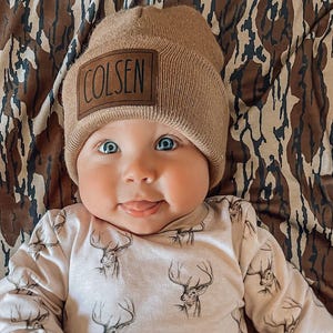 May include: A tan beanie with a brown rectangular patch featuring the name COLSEN in black text, paired with a long-sleeved shirt and pants set in a light beige color, decorated with a repeating black line drawing pattern of deer heads.
