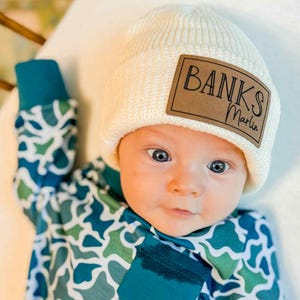 May include: A cream-colored knit beanie featuring a rectangular brown leather patch with the text "BANKS Marlin" in black, paired with a long-sleeved shirt in a blue, green, and white camouflage-style pattern with a solid teal chest pocket.