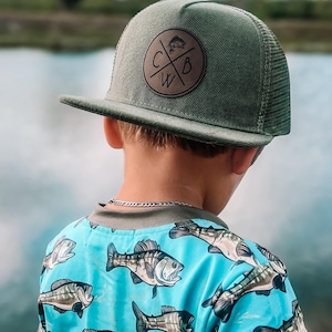 May include: A green trucker hat with a brown leather patch featuring the letters "CWB" and a fish graphic. The child is wearing a blue shirt with a repeating fish pattern. The shirt has gray trim.