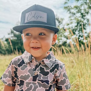May include: A young child wearing a black and grey camouflage polo shirt and a black trucker hat with the name "Colsen" written in white on the front.