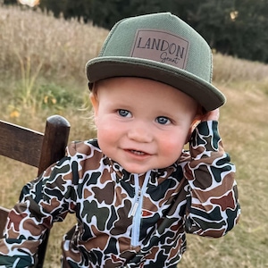 May include: A young child wearing a camouflage print long-sleeve shirt and a green baseball cap with a brown patch that reads "LANDON Grant".
