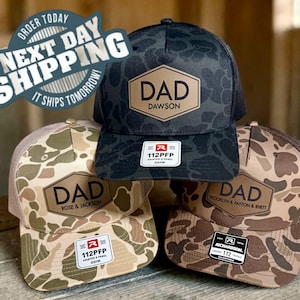 May include: Three camouflage baseball caps with the word "DAD" in a hexagon shape. Each cap has a different name below the word "DAD". The caps are in shades of brown, tan, and black. The caps are on a wooden surface.