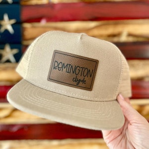 May include: A beige trucker hat with a brown leather patch that says "REMINGTON Clyde" in black lettering.