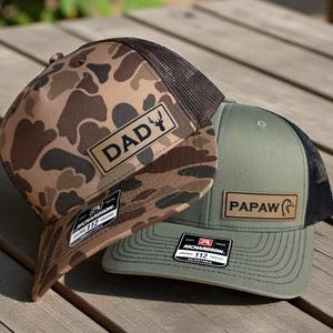 May include: Two Richardson 112 trucker hats. One hat has a camouflage pattern with "DAD" on a leather patch and a brown mesh back. The other is olive green with "PAPAW" on a leather patch and a black mesh back.