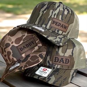 May include: Three camouflage trucker hats stacked on a wooden surface. Each hat has a brown leather patch with the words "PEPAW", "DADDY", and "DAD". The hats have a mesh back and a curved brim.