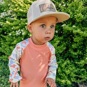 May include: A young child wearing a peach-colored long-sleeved rash guard with a colorful abstract pattern on the sleeves. The child is also wearing a beige mesh trucker hat with a brown patch that says "Colsen" on it.