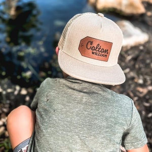 May include: A child wearing a beige mesh trucker hat with a brown leather patch that says "Colton William" in cursive. The child is wearing a green t-shirt and camouflage shorts.