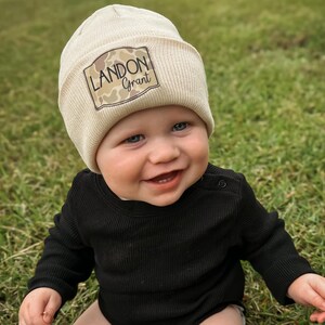 May include: A cream-colored ribbed beanie featuring a rectangular patch with a camouflage pattern and the text LANDON Grant in black lettering, paired with a long-sleeved black ribbed bodysuit with a snap closure at the shoulder neckline.