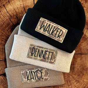 May include: Three knit beanies in black, beige, and tan, each with a rectangular patch. The patches feature names in black lettering against a camouflage pattern. The names are "WALKER", "BENNETT", and "KAYCE".