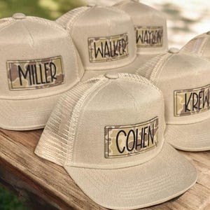May include: A collection of beige trucker hats with mesh backs and curved brims. Each hat features a rectangular patch with a name printed in black over a camouflage pattern. Names include "MILLER", "WALKER", "WAYLON", "KREW", and "COHEN".