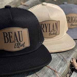 May include: Three personalized trucker hats in black, tan, and gray. Each hat features a leather patch with a name and a second name below it. The black hat reads "BEAU Elliot". The tan hat reads "RYLAN Beckett". The gray hat reads "RIGGS Harrison".