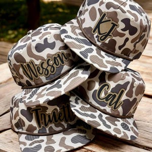 May include: A stack of camouflage print baseball caps with personalized text. The caps feature a brown brim and are decorated with names or initials in a contrasting color. The caps are stacked on a wooden surface.