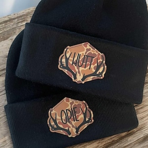 May include: Two black beanies with a folded brim. Each beanie features a brown and tan camouflage patch in a hexagonal shape with deer antlers and the names "HUTT" and "OPIE" in black lettering.