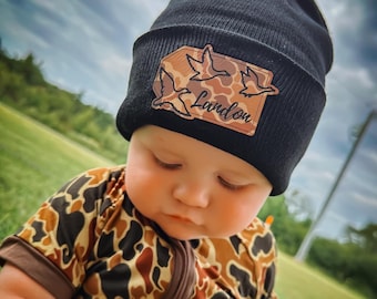 May include: A baby wearing a black beanie with a brown patch featuring a camouflage pattern and the name Landon. The baby is wearing a camouflage onesie.