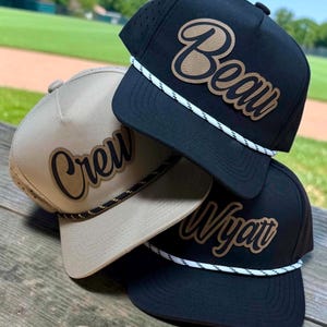 May include: Three baseball caps featuring a flat brim and a rope detail across the base of the crown. Two caps are black with white rope and one is tan with black rope. Each cap displays a custom name in a script font: Beau, Crew, and Wyatt.