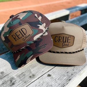 May include: Two baseball caps with rope trim across the brim. One cap features a green, brown, and black camouflage pattern with a tan leather-like patch reading REID William. The other is solid tan with a matching patch reading CRUE David.