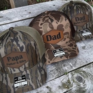 May include: Three camouflage trucker hats with brown leather patches. The patches read "Papa Parker & Riley" and "Dad Parker & Riley". The hats have a mesh back and a curved brim. The hats are adjustable and have a Richardson 112 tag.