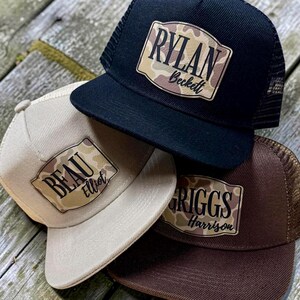 May include: Three trucker hats in black, tan, and brown, each with a personalized patch. The patches feature names like "Rylan," "Beau," and "Griggs" in a camouflage design. The hats are stacked on a wooden surface.