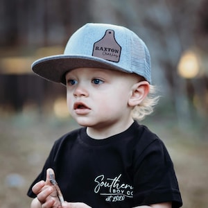 May include: A young child wearing a gray and blue mesh trucker hat with a brown leather patch that says "RAXTON Charles". The child is wearing a black t-shirt with white text that says "Southern Boy Co. est. 2020".