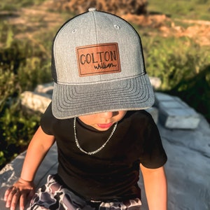 May include: A young person wearing a gray and black trucker hat with a brown leather patch that says "COLTON william". The hat has a black mesh back. The person is wearing a black t-shirt and camouflage pants.